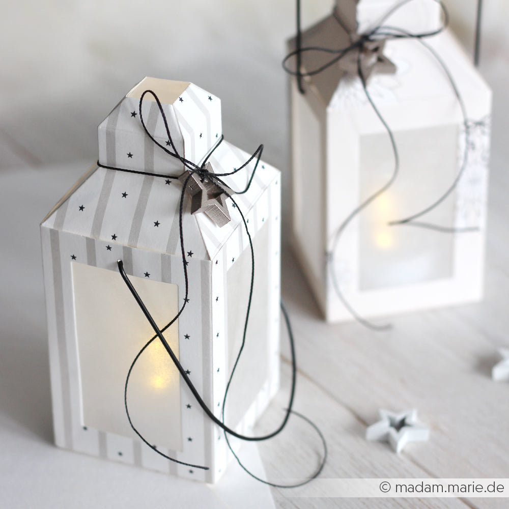 Zwei weiße Papierlaternen mit schwarzen Sternenmustern, die sanft leuchten und mit schwarzen Kordelschleifen verziert sind - hergestellt mit dem Alexandra Renke Laternen Box DieCut System für eine charmante, persönliche Note.