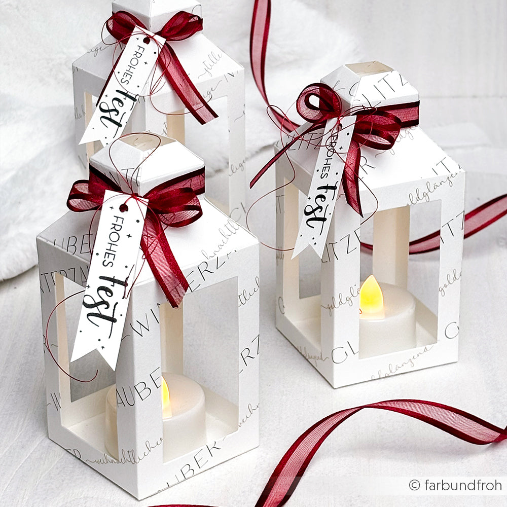 Drei weiße Papierlaternen aus der Alexandra Renke Motivstanze Laternen Box, LED-Kerzen und roten Bändern, jeweils mit der Aufschrift "Frohes Fest".