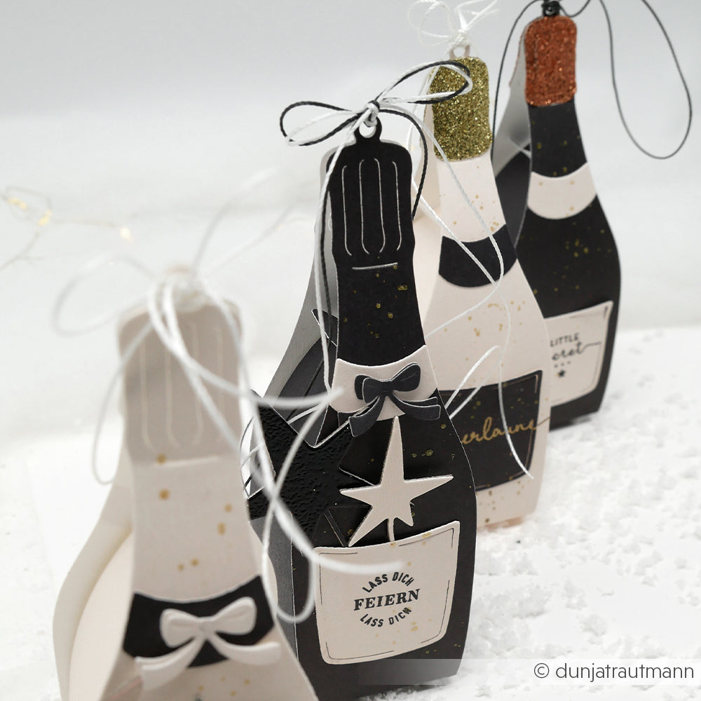 Vier dekorative Sektflaschen aus Papier von Alexandra Renke, hergestellt mit DieCut Systemen und glitzernden Designpapieren, jede mit einer Schleife verziert und in einer Reihe auf einer schneeweißen Fläche angeordnet.