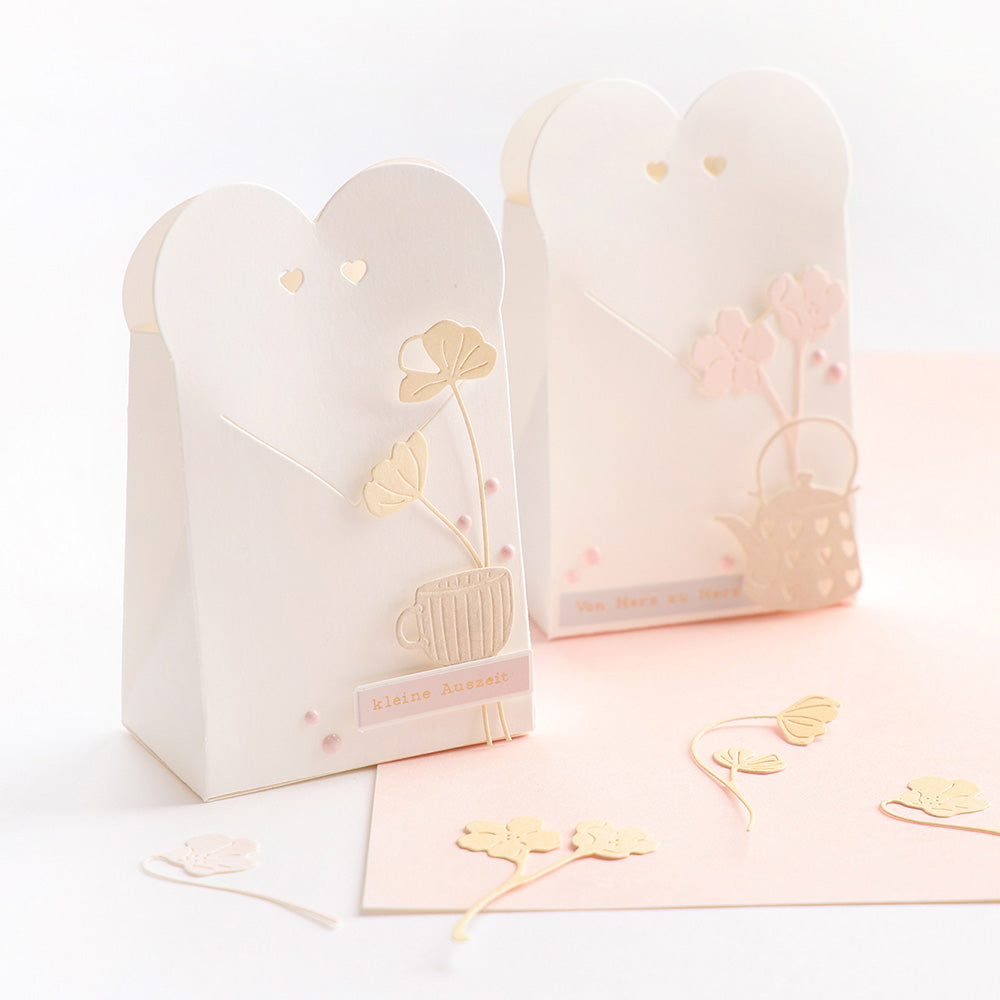 Zwei pastellfarbene Geschenktüten mit floralen Verzierungen und herzförmigen Verschlüssen, gefertigt mit Alexandra Renke Motivstanze Herz-Tüte und eleganten Designpapieren, ruhen auf einer hellrosa Fläche.