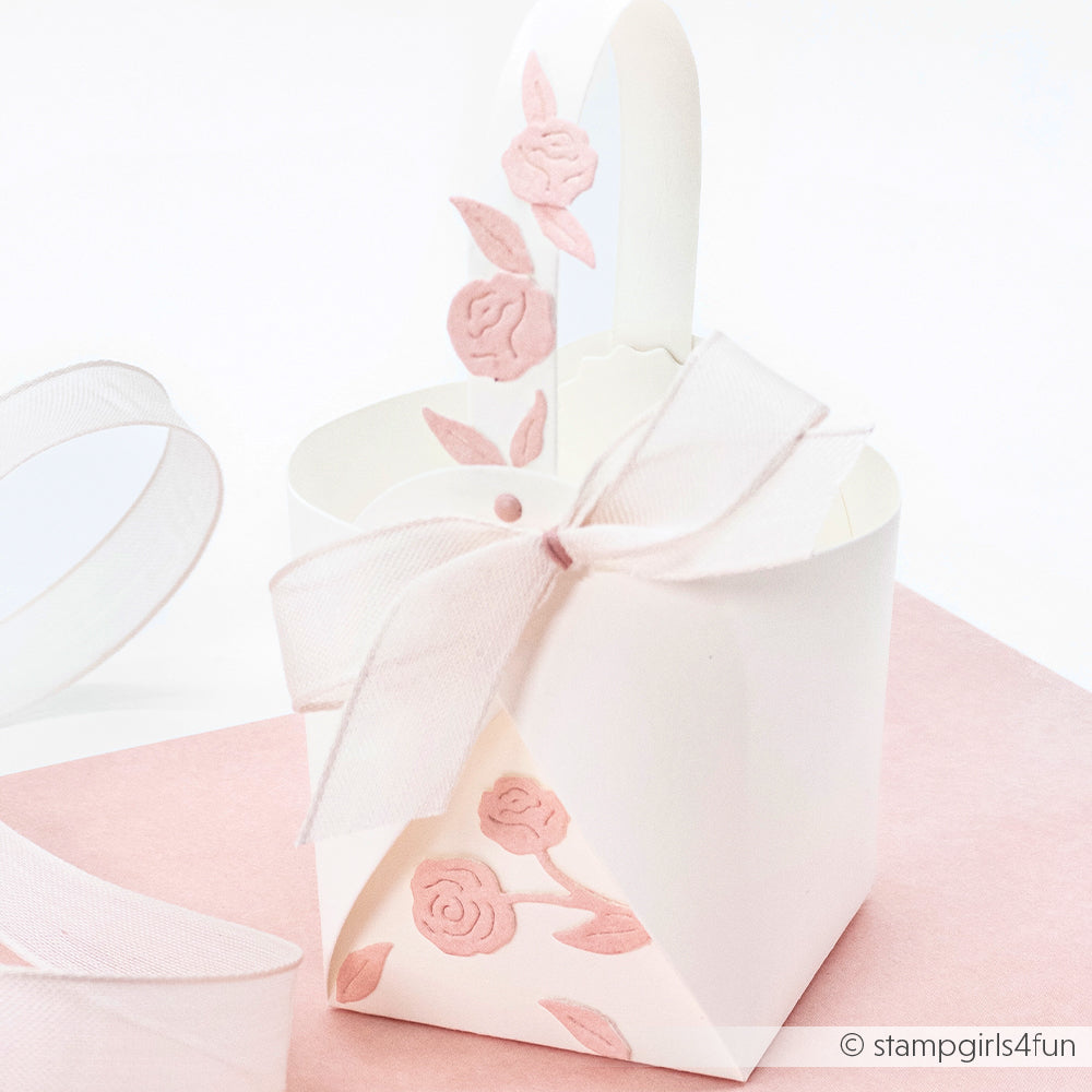 Geschenkkorb aus weißem Papier mit rosafarbenen Blumenmustern, hergestellt mit der Motivstanze Kleines Körbchen von Alexandra Renke und verziert mit einer weißen Schleife, ausgestellt auf einer blassrosa Fläche.