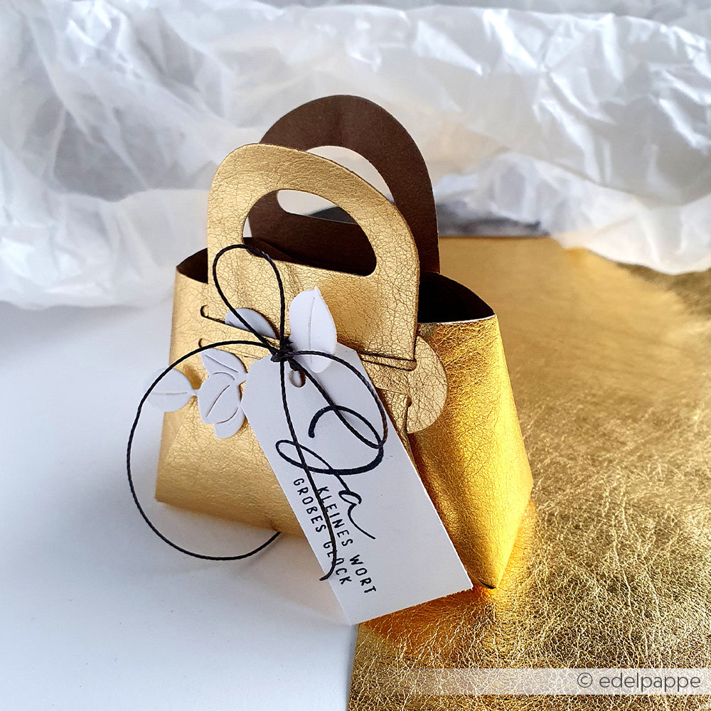 Kleine Geschenktüte aus Goldpapier mit schwarzer Kordel und weißem Anhänger, gefertigt mit Alexandra Renke Motivstanze Steck-Tasche auf Premium Designpapier Stanzen, platziert auf einer gold-weiß strukturierten Oberfläche.