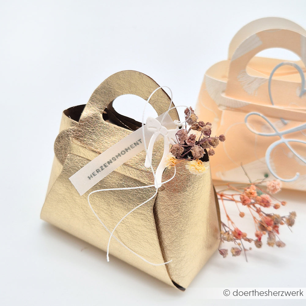Gold- und pfirsichfarbene Geschenktaschen in Handtaschenform von Alexandra Renke, hergestellt aus Designpapier und veredelt mit Motivstanze Steck-Tasche DieCut Systeme, verziert mit Trockenblumen und Anhängern, ausgestellt auf weißem Hintergrund.