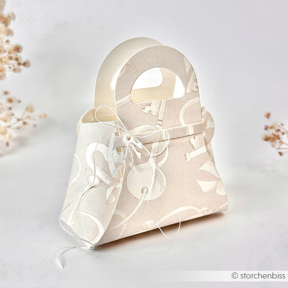 Elegante, weiße Geschenkschachtel in Form einer Handtasche von Alexandra Renke, hergestellt aus Designpapier mit Blumenmustern und einer Schleife vor einem weichen, neutralen Hintergrund.