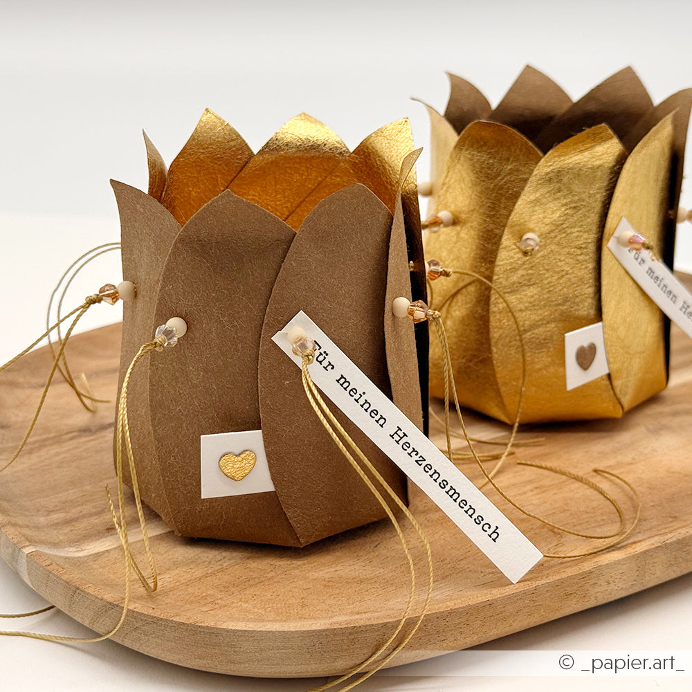Auf einem Holztablett stehen zwei goldene und braune lotusförmige Geschenkschachteln von Alexandra Renke aus Motivstanze Blütenkörbchen mit Herzanhängern und Goldkordel.