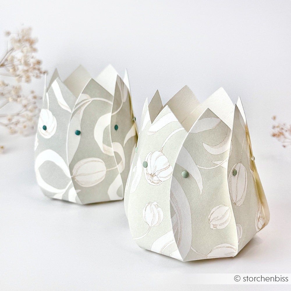Zwei pastellfarbene, tulpenförmige Papierlaternen von Alexandra Renke aus Designpapier stehen auf einer weißen Fläche mit zarten Trockenblumen daneben.