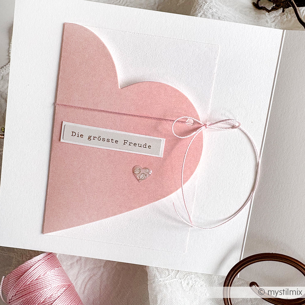 Eine rosafarbene Herzkarte von Alexandra Renke, hergestellt mit der Motivstanze Herzenskarte, mit deutschem Text, einer Schleife und einem kleinen silbernen Herzanhänger auf weißem Designpapier.