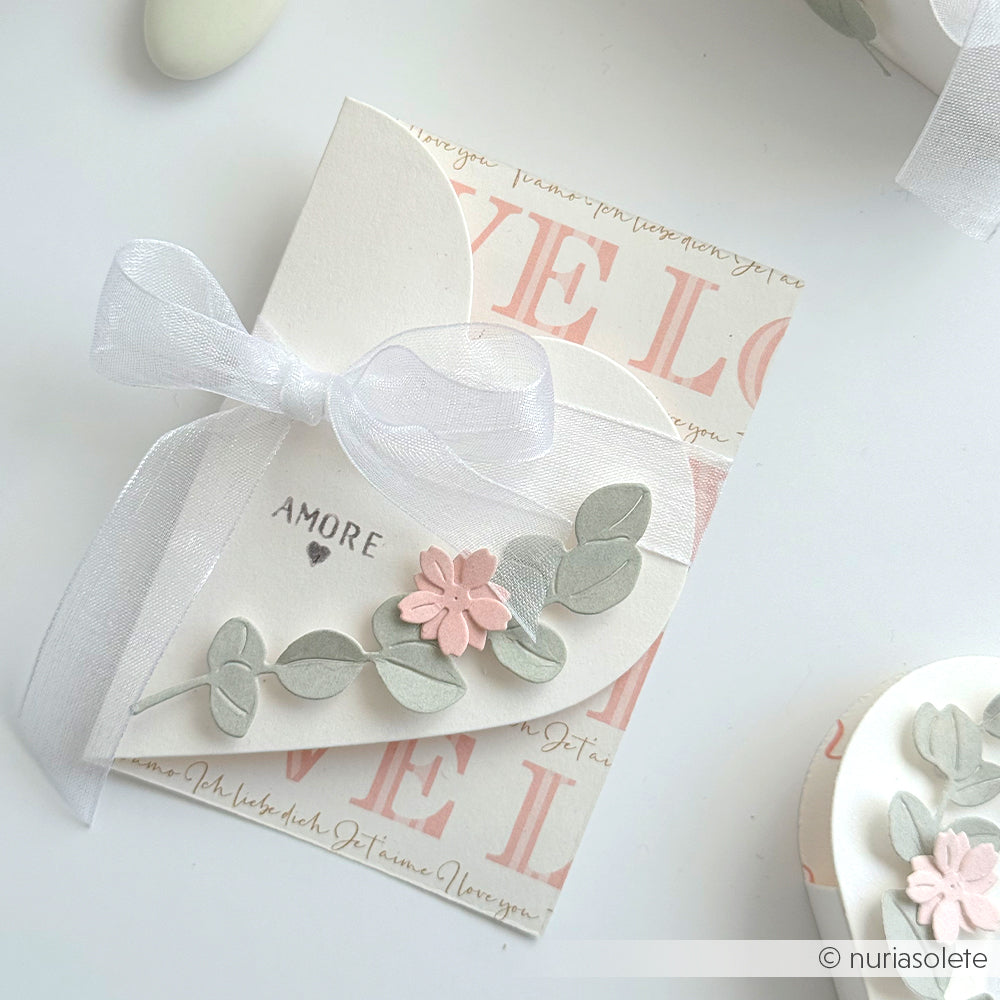 Handgefertigte Karte mit AMORE-Text, floralen Akzenten und weißem Band, hergestellt mit Alexandra Renke Motivstanze Herzenskarte und Designpapieren, vor einem romantischen Hintergrund.