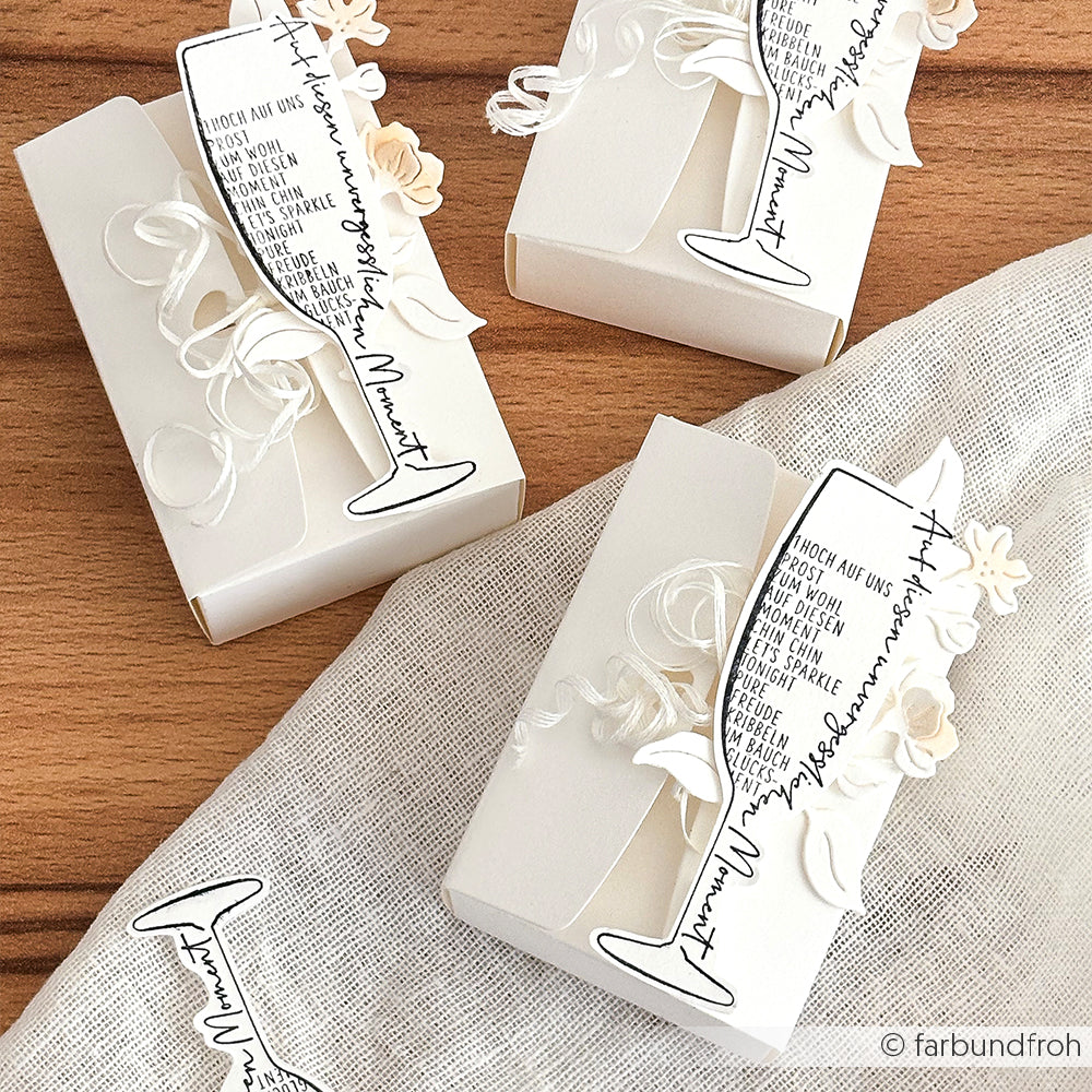 Drei elegante weiße Geschenkboxen von Alexandra Renke, die mit Stanzen für Designpapier gefertigt und mit dekorativen Sektgläsern und Blumen aus Papier verziert sind, ruhen anmutig auf einer strukturierten Stoffoberfläche.