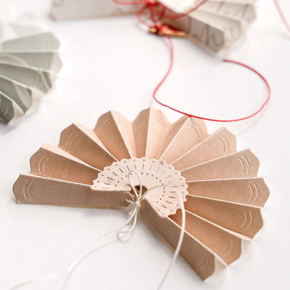 Ein Fächerschmuck aus Alexandra Renke Designpapieren und einer Motivstanze Faltfächer klein ruht auf einer weißen Fläche, umgeben von weiteren Fächern und roter Schnur.
