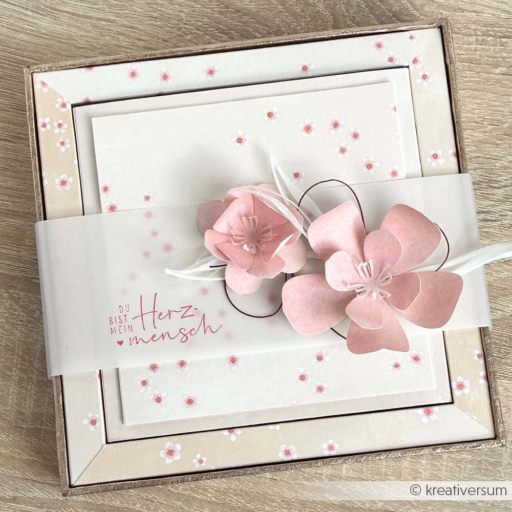 Eine quadratische Blumenkarte von Alexandra Renke mit rosa Papierblumen, die mit Designpapieren und Motivstanze 3D Kirschblüte hergestellt wurden, sowie dem deutschen Text "Du bist mein Herzmensch" auf einem Pergamentband.