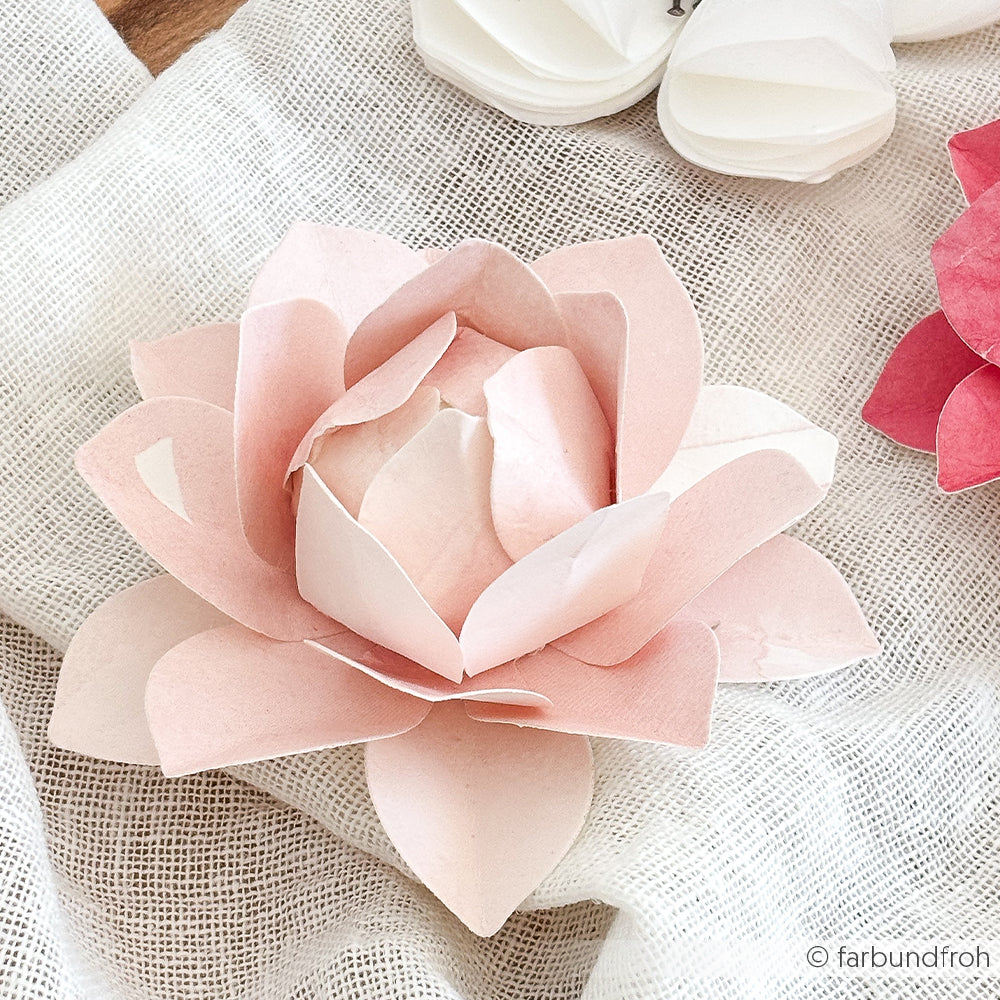 Rosafarbene Lotosblüte aus Papier mit übereinander liegenden Blütenblättern, hergestellt mit Alexandra Renke Designpapieren und einer Motivstanze, auf einem strukturierten weißen Stoffhintergrund.