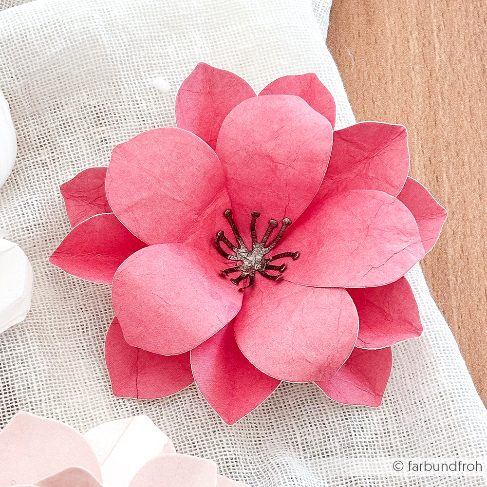 Alexandra Renke rosa 3D-Kirschblüte aus Papier mit geschichteten Blütenblättern und detaillierten Staubgefäßen, hergestellt aus Designpapier, auf einer weißen, strukturierten Stoffoberfläche.