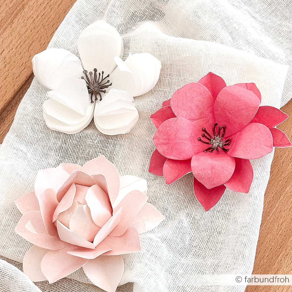 Drei zarte 3D-Kirschblüten aus Papier in Weiß und Rosa, hergestellt mit Alexandra Renke Designpapieren, werden auf einem weißen Tuch auf einer Holzfläche präsentiert.