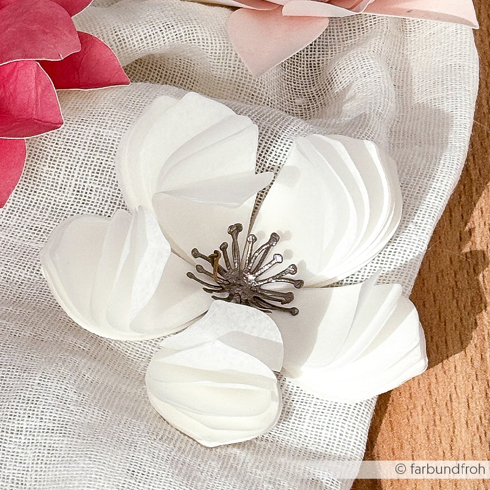 Eine weiße Papierblume mit einer detaillierten Mitte, die mit Alexandra Renke Designpapieren hergestellt wurde, ruht auf weißem Stoff neben rosa 3D-Kirschblütenblättern.