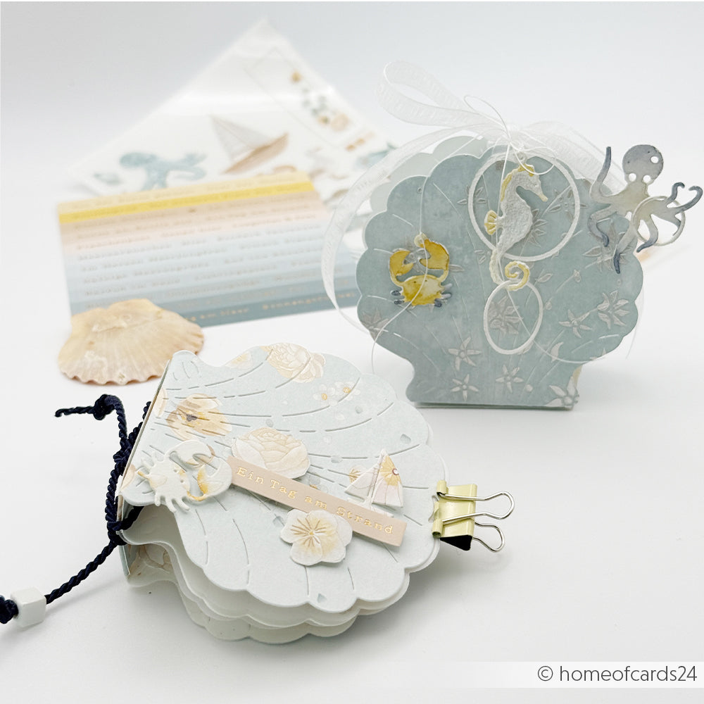 Die handgefertigten Karten in Muschelform von Alexandra Renke sind mit Meeresmotiven verziert, die aus Designpapier gefertigt und mit der Motivstanze „Muschel Box“ verziert sind – alles vor einem weißen Hintergrund.