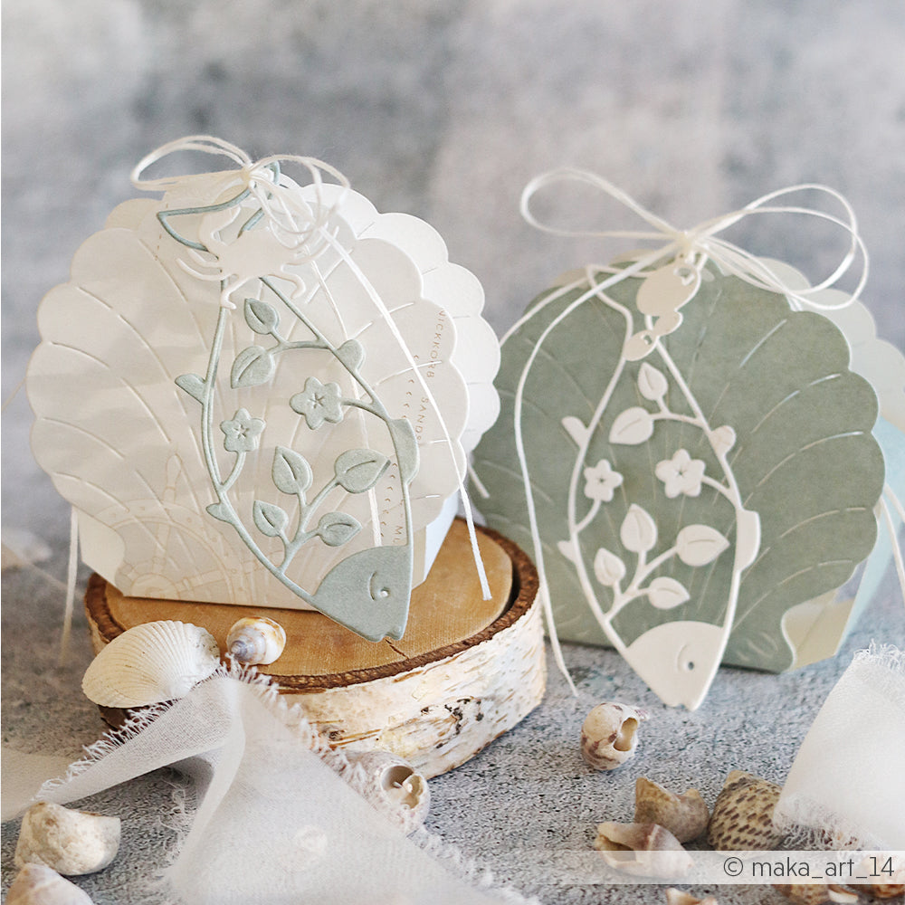 Zwei Karten in Form von Jakobsmuscheln von Alexandra Renke, gefertigt aus Designpapier und Stanzteilen, mit Fisch- und Blattmotiven. Sie sind mit einer weißen Schnur zusammengebunden und werden zwischen Muscheln präsentiert.