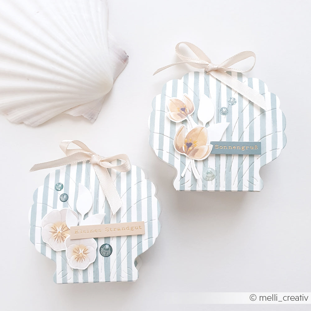 Zwei muschelförmige Geschenkboxen mit Schleifen, Streifenmustern und floralen Verzierungen – gefertigt aus Designpapieren von Alexandra Renke und mit dem Stanzsystem „Muschel Box“ von Motivstanze – sind neben einer weißen Muschel ausgestellt.