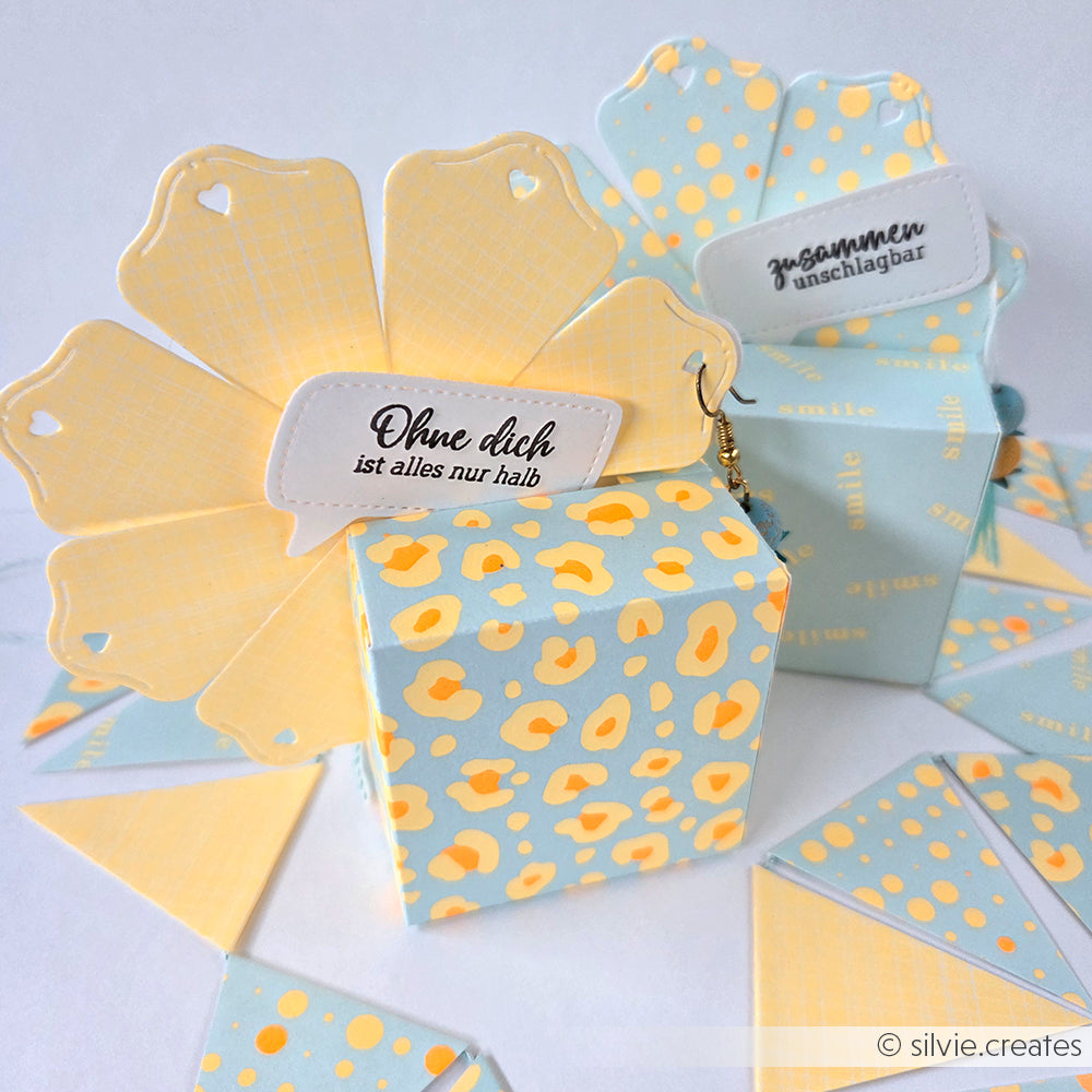 Pastellblaue und gelbe Geschenkboxen mit Blumenkarten und deutschem Text, verziert mit passenden Papiergirlanden, zeigen den kreativen Einsatz von Stanzsystemen und der Motivstanze „Fächerbox“ in Kombination mit passenden Designpapieren.
