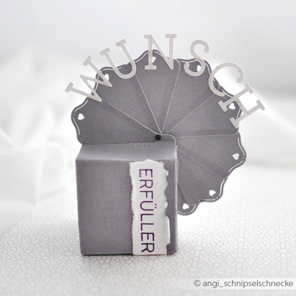 Graue Geschenkbox mit einem dekorativen Fächer, gefertigt aus Alexandra Renke Designpapieren und mit der Alexandra Renke Motivstanze „Fächerbox“, auf deren weißer, strukturierter Oberfläche die deutschen Wörter „WUNSCH“ und „ERFÜLLER“ zu sehen sind.