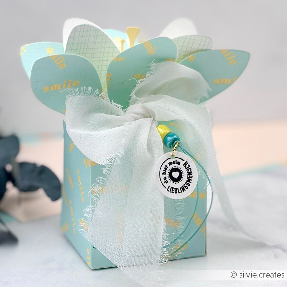 Hellblaue Geschenkbox mit Blumenmuster und „Smile“-Aufdruck, weißem, durchsichtigem Band und einem runden Anhänger an der Vorderseite. Hergestellt aus erlesenen Designpapieren und mit einer Motivstanze aus dem Blumenbox-Stanzsystem verziert, was ihr zusätzliche Eleganz verleiht.