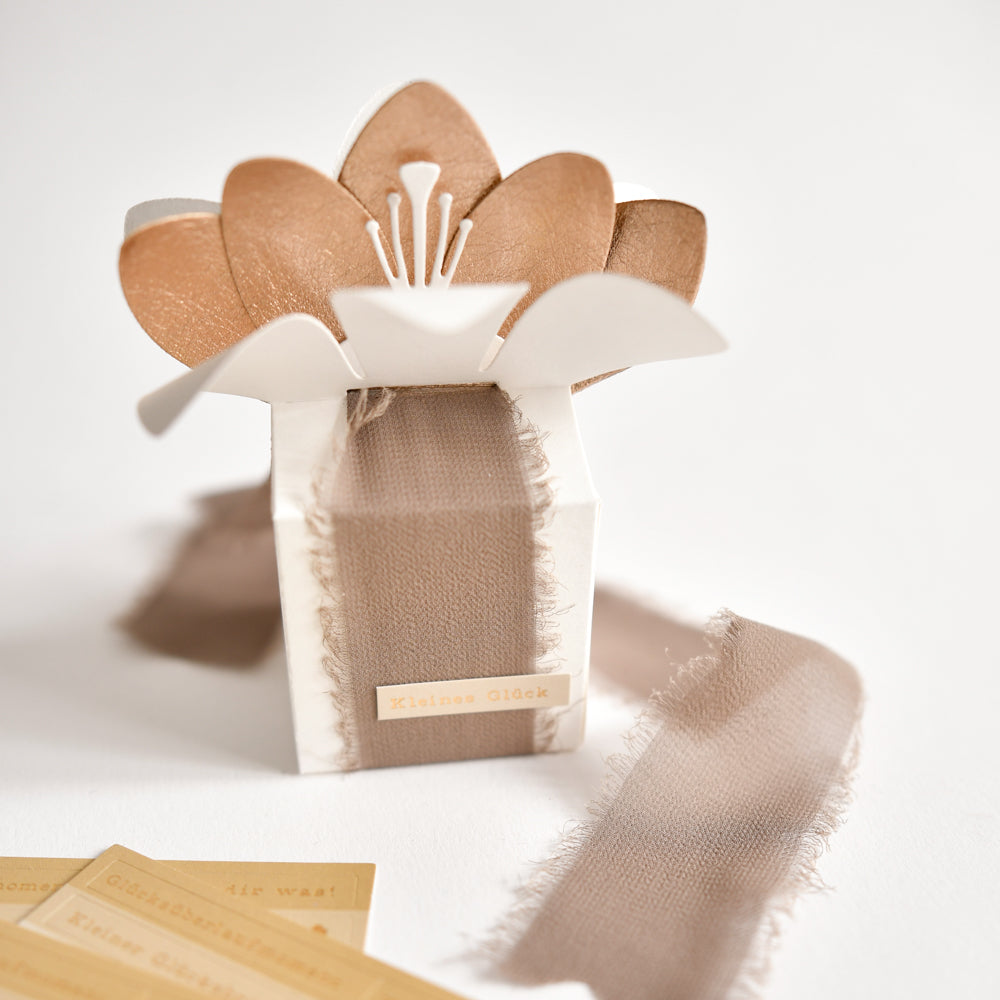 Eine kleine Schachtel von Alexandra Renke, verziert mit einem Stoffband und einem dekorativen, blumenförmigen Deckel, der aus Designpapieren gefertigt und mit der Motivstanze „Blumenbox“ von DieCut Systeme verziert wurde, steht elegant auf einem weißen Hintergrund.