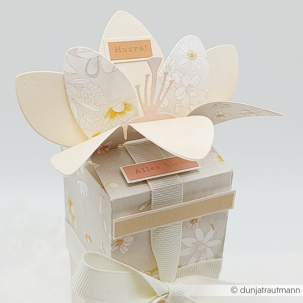 Eine blumenförmige Karte auf einer Geschenkbox, gestaltet mit eleganten Designpapieren von Alexandra Renke, verziert mit deutschen Grußtexten und Blumenmustern und mit einem Band verziert – perfekt gestaltet mit der Motivstanze „Blumenbox“ von Stanzsysteme.