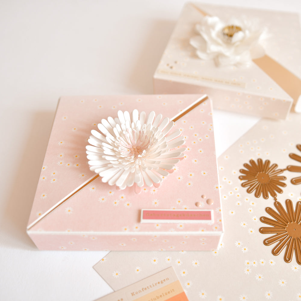 Rosa Geschenkbox mit einem weißen Blumenverzierung, die mit dem „3D Pompom Blume DieCut System“ von Alexandra Renke gefertigt wurde, präsentiert neben goldenen und weißen Blumenausschnitten auf Papier, die mit der „Motivstanze“ von Alexandra Renke hergestellt wurden.