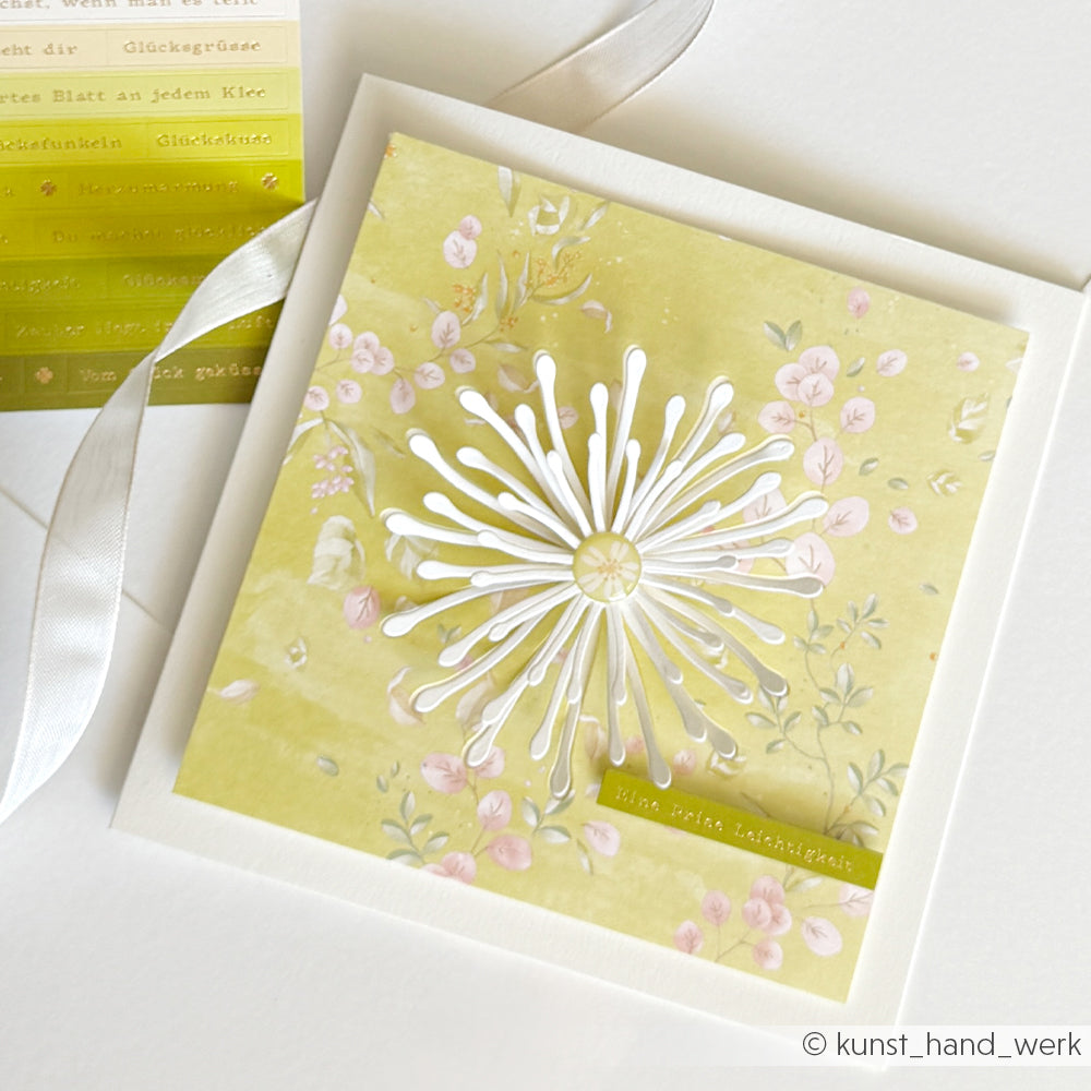 Quadratische Blumenkarte von Alexandra Renke mit einem weißen, ausgestanzten Löwenzahn in einem bezaubernden 3D-Pusteblumen-Design auf leuchtend gelbem Hintergrund, daneben ein Band und ein Etikettenbogen – ein perfektes Beispiel für Kreativität mit Motivstanze und DieCut Systeme.