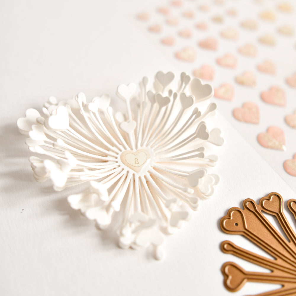 Ein Herzausschnitt aus weißem Papier mit strahlenförmig angeordneten Herzformen, gestaltet mit Designpapieren von Alexandra Renke und der Stanze „Motivstanze 3D Herzchen Blume“, präsentiert zusammen mit einem goldenen Ausschnitt und pastellfarbenen Herzaufklebern auf weißem Untergrund – eine perfekte Kombination für Stanzbegeisterte.