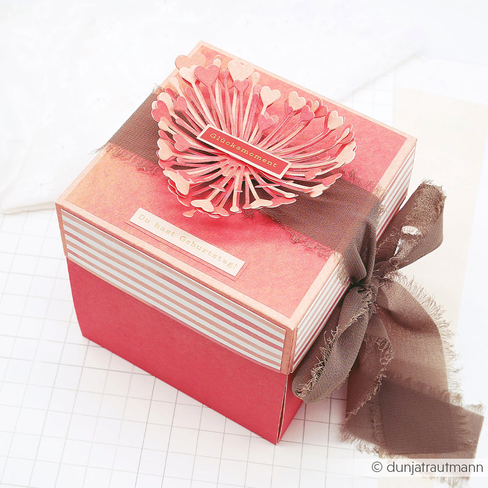 Rote Geschenkbox mit einer herzförmigen Papierverzierung, die mit der „Motivstanze 3D Herzchen Blume“ von Alexandra Renke gestaltet wurde, verziert mit einem braunen Band und einem deutschen Geburtstagsgruß oben drauf.