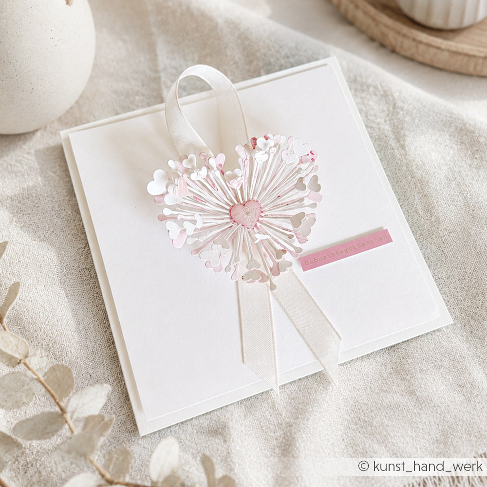 Quadratische weiße Karte von Alexandra Renke mit einem rosa, herzförmigen Löwenzahn-Motiv, das mit einer 3D-Stanze „Herzchen Blume“ ausgestanzt, mit einem zarten Band verziert und auf beigem Stoff angebracht wurde.
