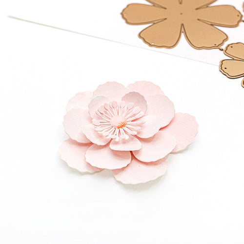 Eine rosa Papierblume mit überlappenden Blütenblättern, die mit den „Motivstanze“-Stanzformen von Alexandra Renke gefertigt wurde, befindet sich neben braunen, blumenförmigen Ausschnitten auf einem weißen Untergrund und bringt das zarte 3D-Design „Wolkenrose“ besonders gut zur Geltung.