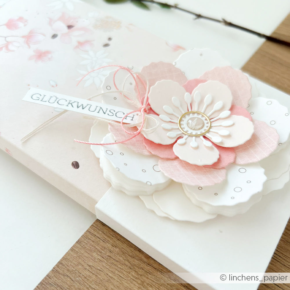 Handgefertigte Karte mit einer mehrschichtigen rosa-weißen Blume, die mit einer Motivstanze von Alexandra Renke gestaltet wurde, verziert mit einem „Glückwunsch“-Anhänger und zarten 3D-Wolkenrose-Details, präsentiert auf einem hellen Holzuntergrund.