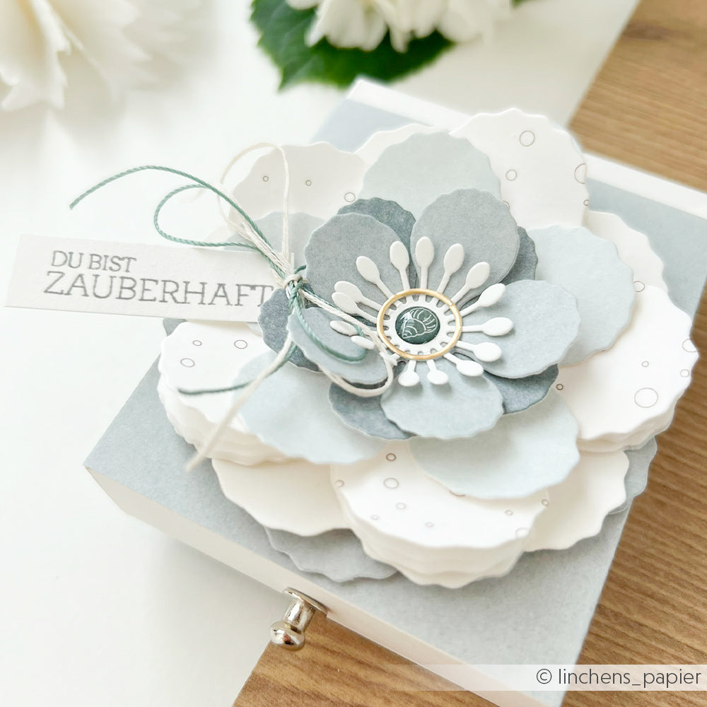 Eine dekorative Papierblume, gefertigt aus eleganten Alexandra Renke Designpapieren und mit der Alexandra Renke Motivstanze 3D Wolkenrose aus der Premium-Reihe „Stanzsysteme“ geformt, ziert wunderschön den Deckel einer hellen Geschenkbox, die mit einem Anhänger mit der Aufschrift „Du bist zauberhaft“ verziert ist.