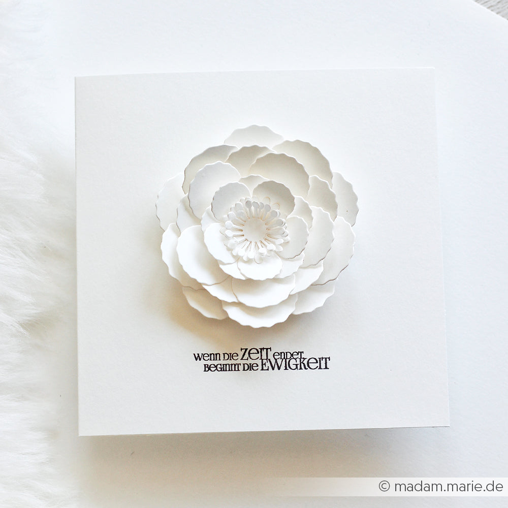 Eine weiße Papierblume, gefertigt mit der 3D-Stanzform „Motivstanze 3D Wolkenrose“ von Alexandra Renke, auf einer quadratischen Karte mit deutschem Text darunter, präsentiert vor weißem Hintergrund.