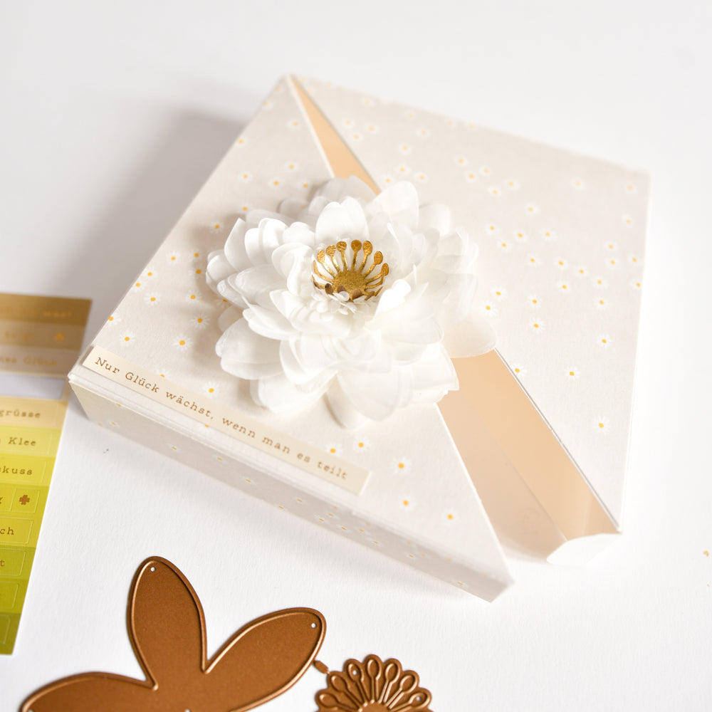 Quadratischer Umschlag, verziert mit einer weißen Papierblume, die mit der Alexandra Renke Motivstanze „3D Sternensee Rose“ gefertigt wurde. Die Blume weist ein gepunktetes Muster auf und wird durch goldene Aufkleber in der Nähe akzentuiert. Ideal für kreative Projekte mit den Stanzsystemen von Alexandra Renke und hochwertigen Designpapieren.