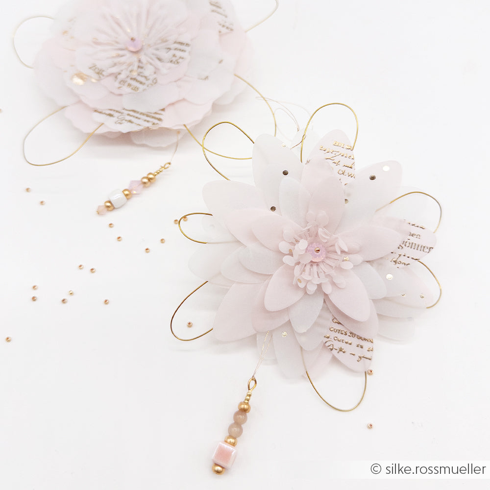 Zarte, mehrschichtige rosa Blumen mit goldenen Drahtschlaufen, Perlen und Textdetails auf weißem Hintergrund werden mithilfe von Designpapieren von Alexandra Renke und dem Stanzsystem „Motivstanze 3D Sternensee Rose“ wunderschön gestaltet.