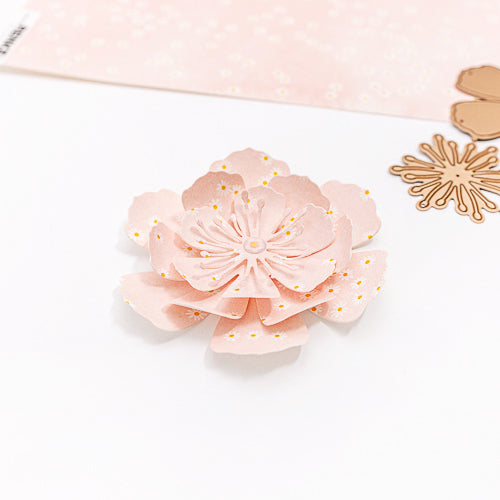 Eine blassrosa Papierblume mit goldenen Punkten, die mit einer 3D-Motivstanze „Pfingstmond Blume“ von Alexandra Renke gefertigt wurde, ist auf einem weißen Untergrund neben verschiedenen Bastelutensilien und Stanzformen zu sehen.
