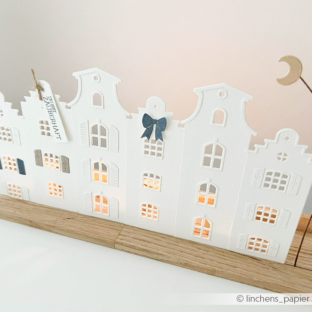 Aus weißem Papier ausgeschnittene Gebäude mit beleuchteten Fenstern auf einem Holzsockel, gefertigt aus hochwertigen Designpapieren und verziert mit kleinen Schleifen und Anhängern. Hergestellt mit der Motivstanze „Häuserbox“ von Stanzsysteme für präzise Details.