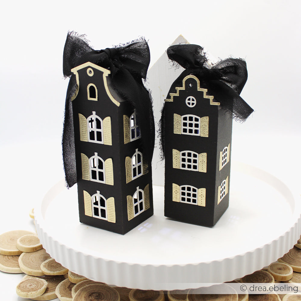 Zwei schwarze, hausförmige Geschenkboxen, die mit der Motivstanze „Häuserbox“ gefertigt und mit großen schwarzen Schleifen verziert wurden, weisen zarte Fenster auf und sind elegant auf einem weißen Teller arrangiert.