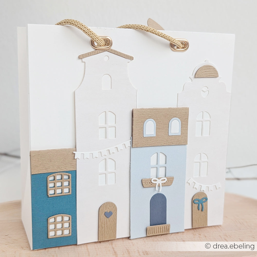 Geschenktüte mit blauen, weißen und beigen Häusern, gefertigt aus Designpapieren und verziert mit ausgestanzten Fenstern und Türen, die mit der Motivstanze „Häuserbox“ aus dem Stanzsystem hergestellt wurden. Mit eleganten goldenen Tragegriffen versehen.