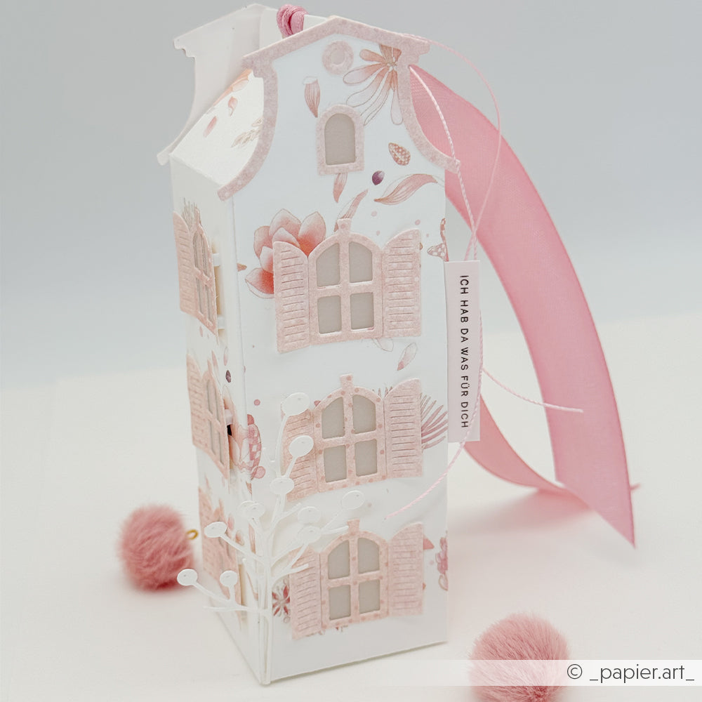 Hohe, rosa-weiße Geschenkbox in Form eines Papierhauses, gefertigt mit der Motivstanze „Häuserbox“ und verziert mit eleganten Designpapieren, abgerundet durch ein großes Band und florale Akzente sowie zarte rosa Pompons.