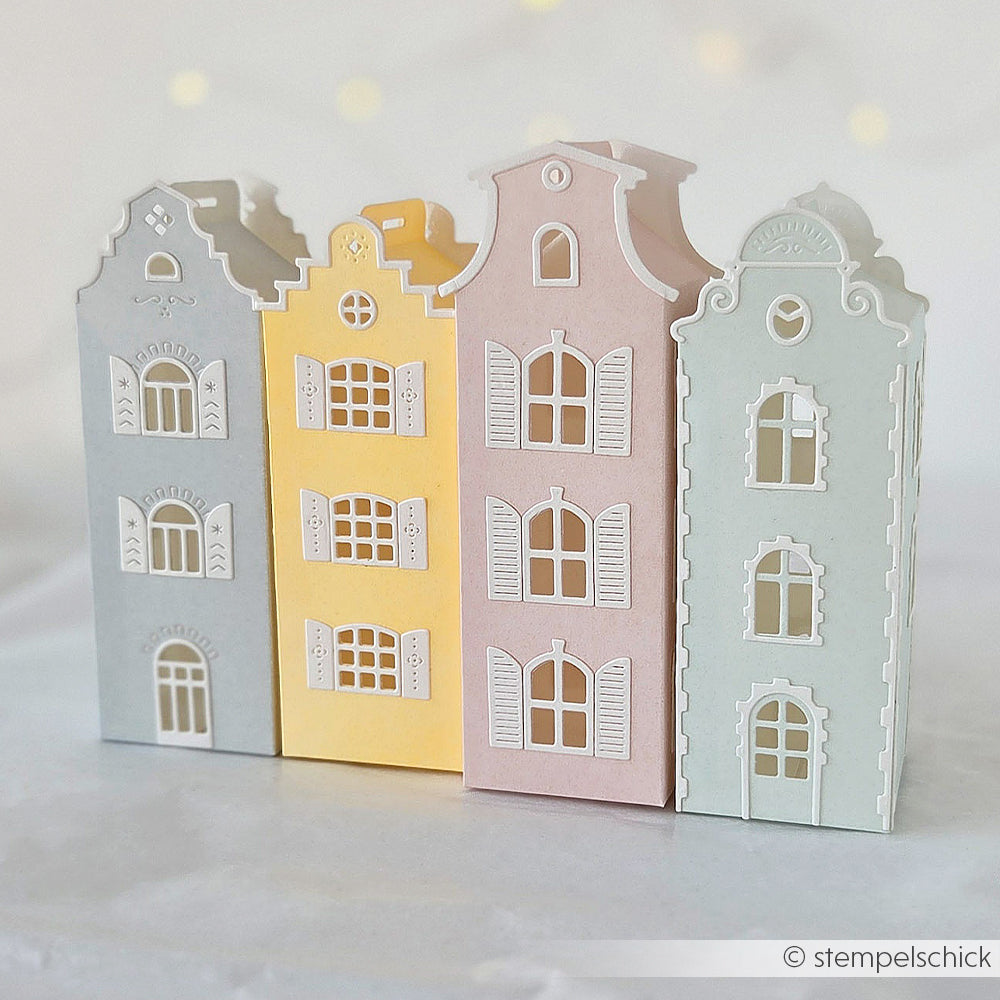 Drei pastellfarbene Papierhäuschen aus Designpapier mit verzierten Fenstern stehen nebeneinander auf einer hellen Unterlage.