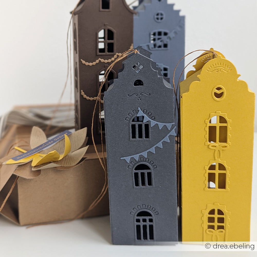 Drei Papierlaternen in Form von Häusern, gefertigt aus edlen Designpapieren in Gelb, Schwarz und Grau, stehen neben einer braunen Schachtel – und zeigen damit perfekt die kreativen Möglichkeiten, die die Motivstanze „Häuserbox“ und andere innovative Stanzsysteme bieten.