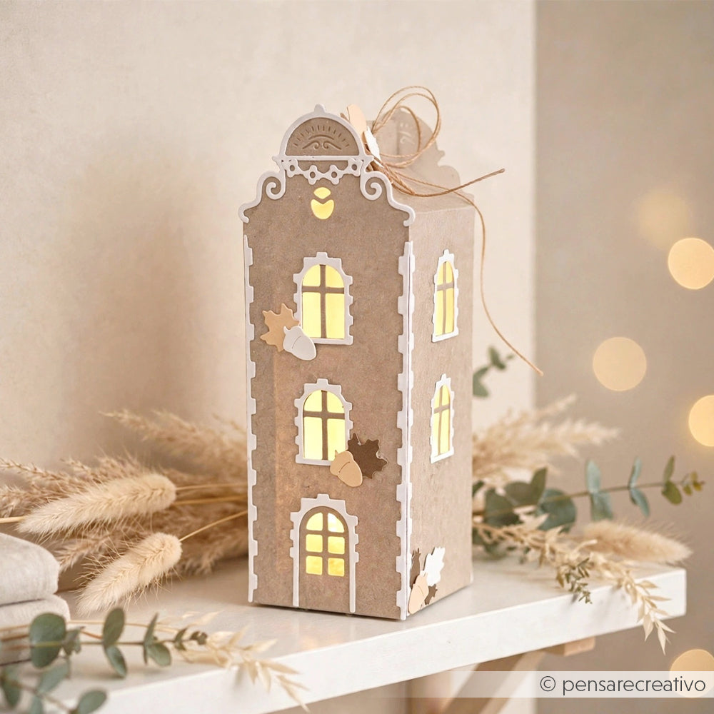 Eine dekorative Hauslaterne aus Pappe, die aus Designpapieren gefertigt und mit Stanzsystemen verziert wurde, steht auf einem Regal; ihre beleuchteten Fenster strahlen warm inmitten von Herbstblättern und einer Schnurschleife oben drauf.