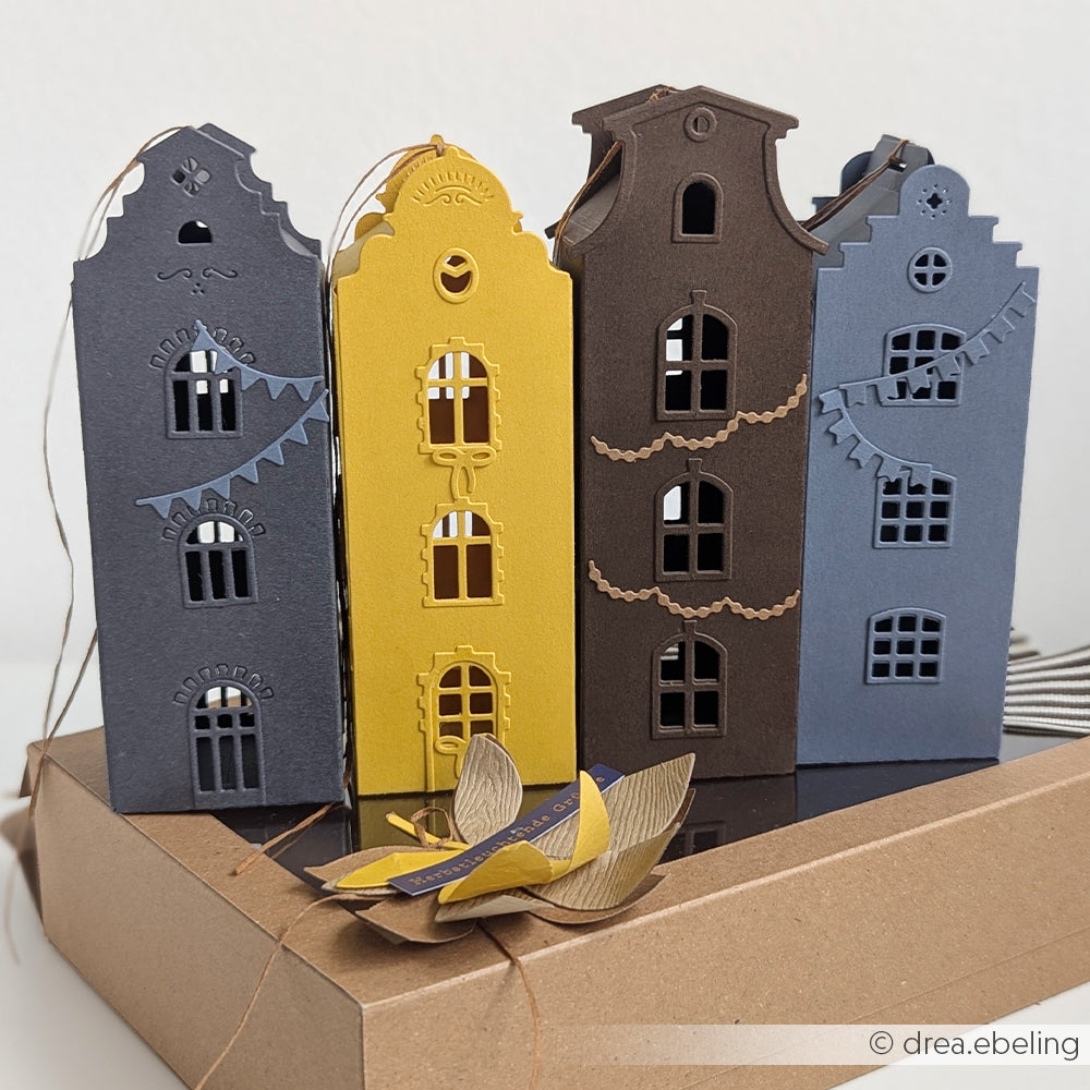 Drei dekorative Papierhäuschen in Blau, Gelb und Braun – gefertigt aus hochwertigen Designpapieren – stehen auf einer Schachtel, die mit der Motivstanze „Häuserbox 3“ von DieCut Systeme gestaltet wurde, und davor sind hübsche Papierblätter angeordnet.
