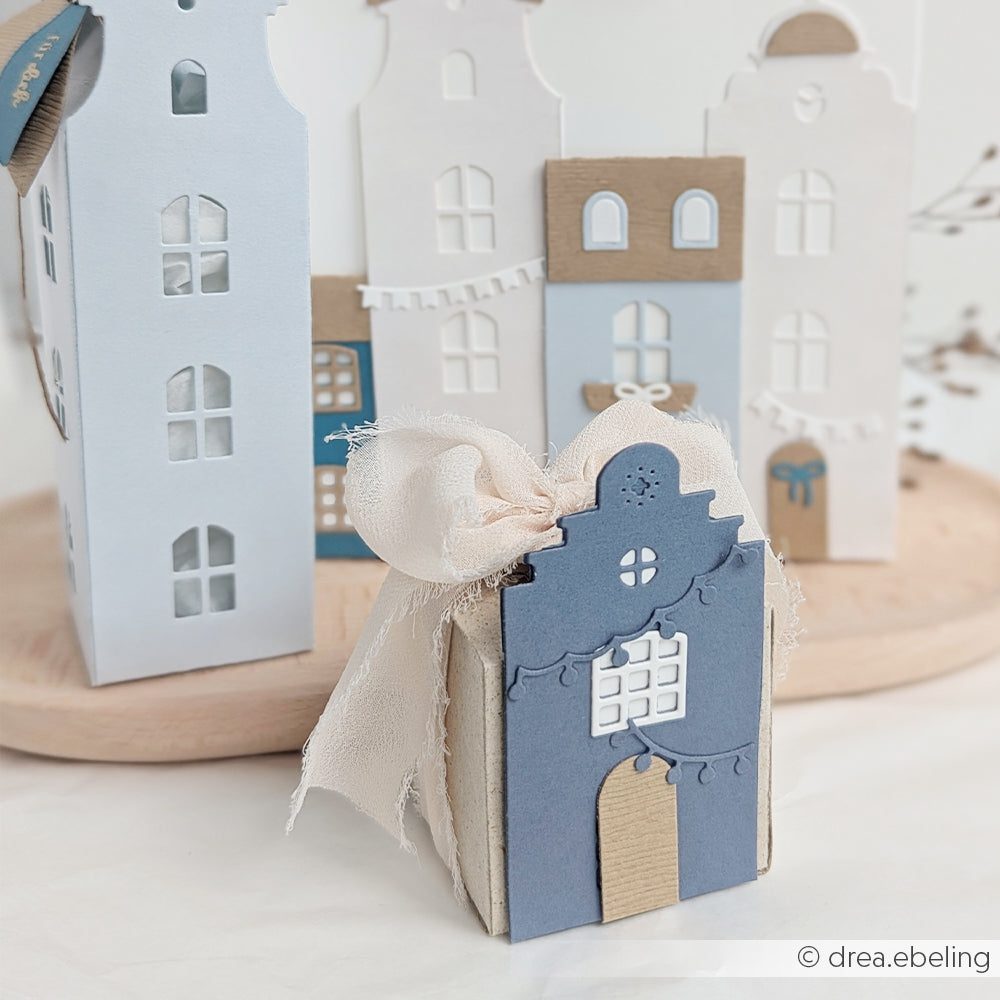 Eine kleine, blau-beige, hausförmige Geschenkbox mit einer Schleife, gefertigt mit der Motivstanze „Häuserbox“ und stilvollem Designpapier, umgeben von ausgeschnittenen Hausverzierungen.