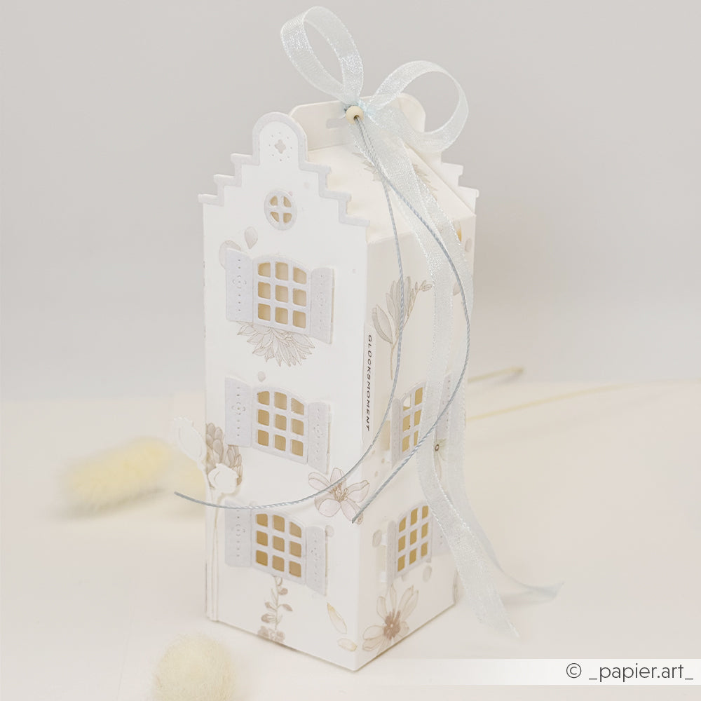 Weiße, hausförmige Geschenkbox, gefertigt mit dem Stanzsystem „Motivstanze Häuserbox 3“, verziert mit floralen Details, Fenstern und einer Schleife aus transparentem Band oben drauf – perfekt, um Ihre Lieblings-Designpapiere in Szene zu setzen.
