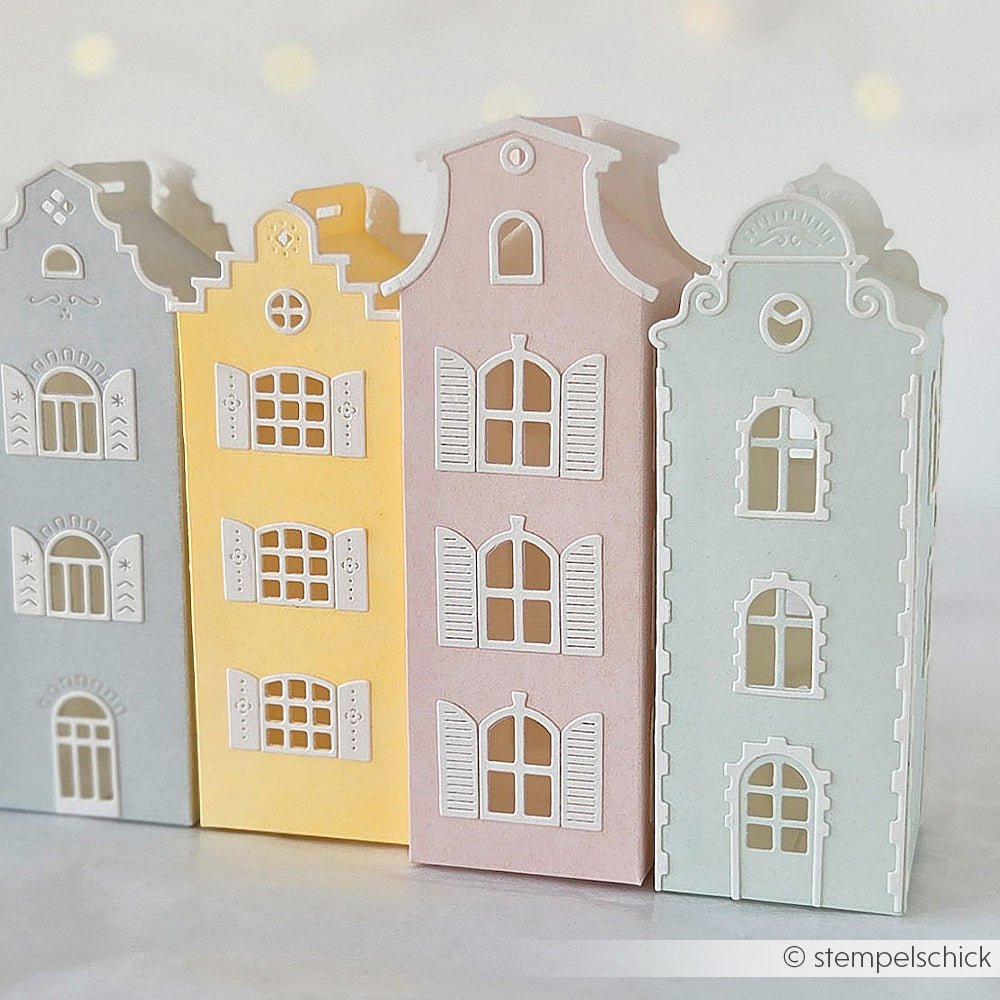Vier pastellfarbene Papierhäuschen, gefertigt aus hochwertigen Designpapieren und mit kunstvoll gestalteten Fenstern und Türen versehen, sind nebeneinander auf einem hellen Hintergrund angeordnet. Jedes dieser bezaubernden Häuschen zeigt, welche filigranen Details mit unseren Motivstanze-Häuserbox-Stanzsystemen möglich sind.